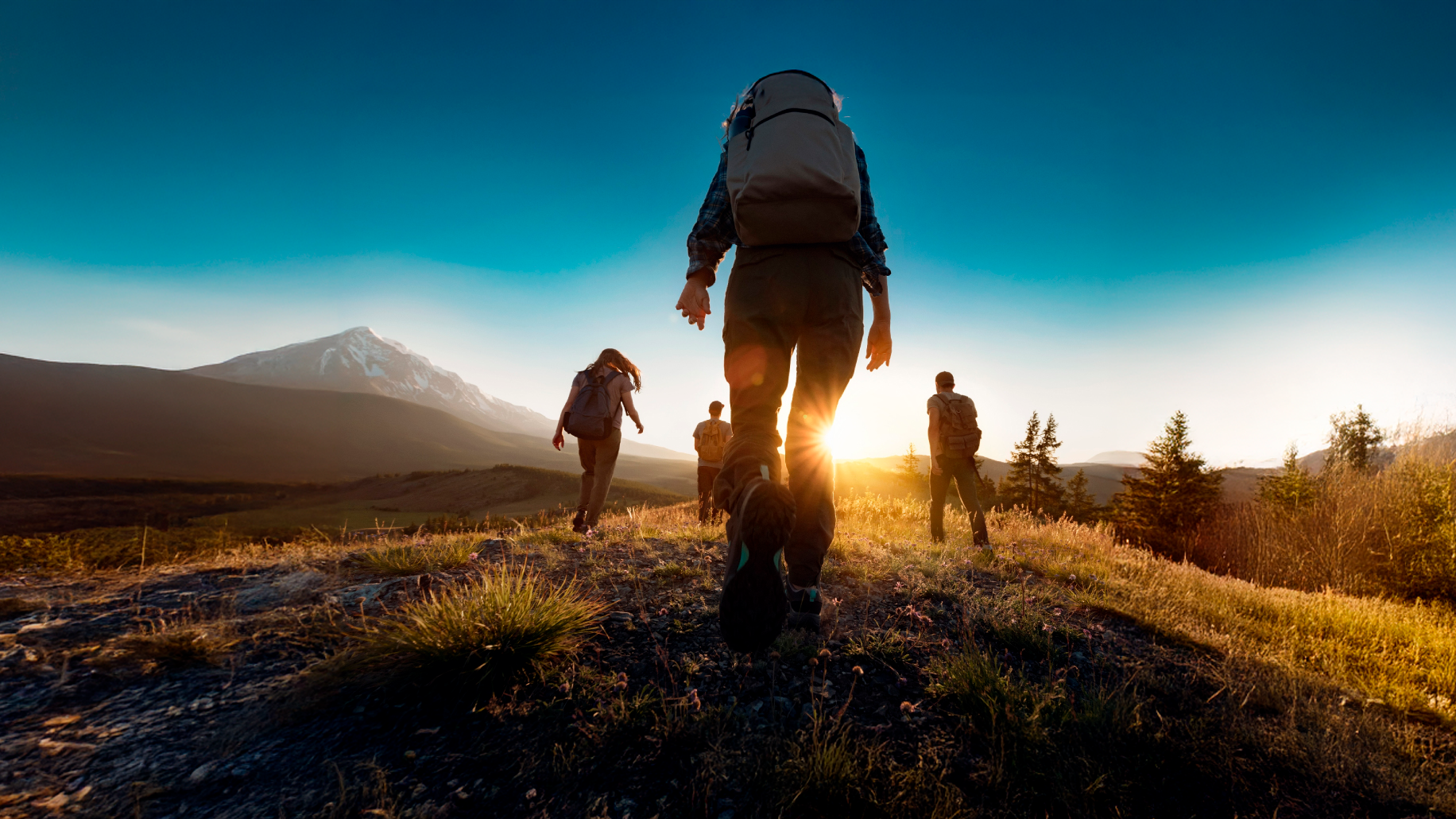 Hikers with backpacks walking on a trail towards the sun over mountains.